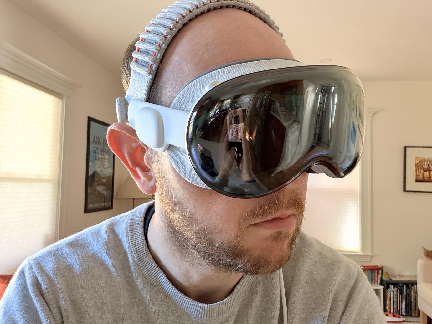 A man wearing a white virtual reality headset with a reflective visor in a well-lit room with framed pictures on the walls.