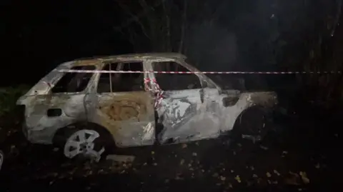 Councillor Craig Monks A burnt out white Range Rover in the dark with red and white tape around it