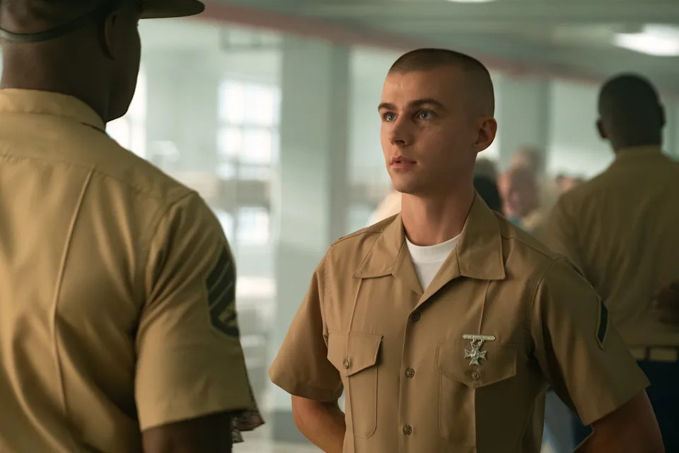 A young person in a military uniform stands facing an officer in a training facility, appearing attentive and serious
