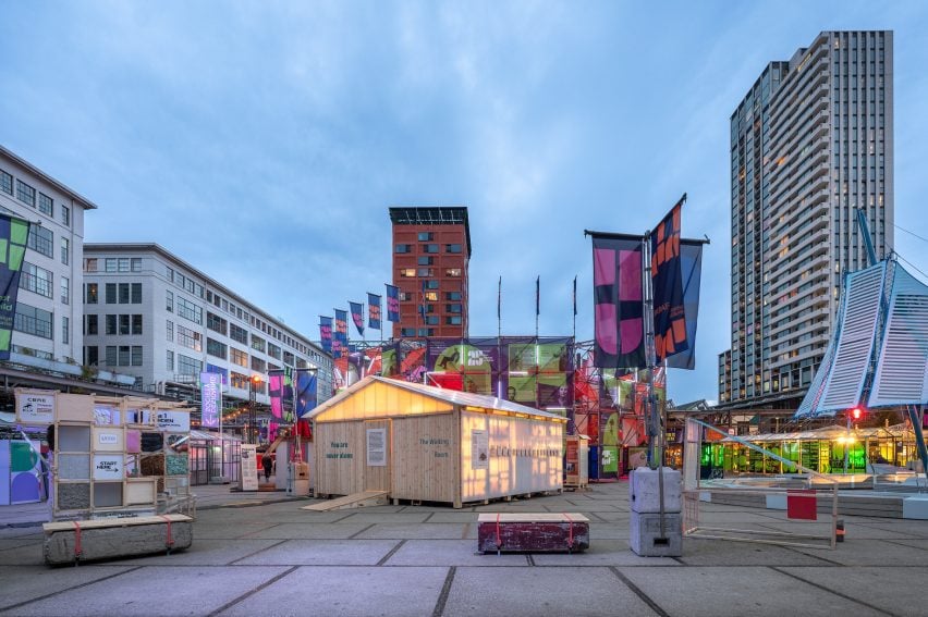 Photo of the Waiting Room pavilion lit up from within at dusk at Dutch Design Week 2025, surrounded by colourful flags and installations