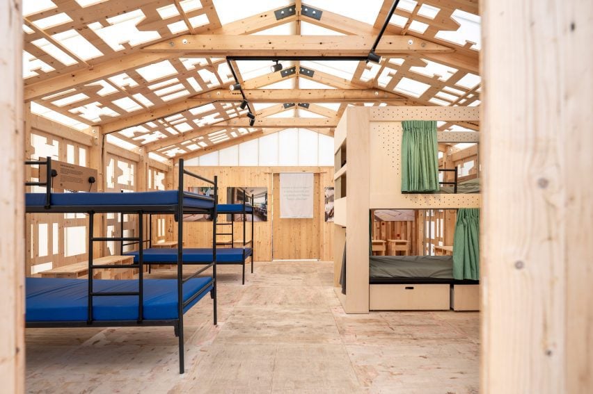 Photo looking into the interior of The Waiting Room pavilion showing spartan bunk beds made of slender black tubular metal on the left-hand side and blonde wood bunk-bed cabins on the right