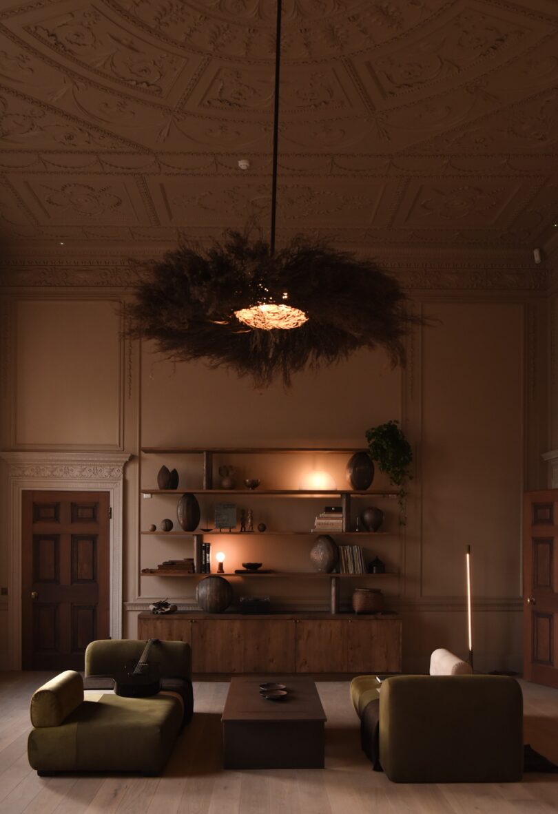 A dimly lit living room with ornate ceiling, dark wood furniture, olive green armchairs, a low coffee table, shelves with pottery, and a large decorative chandelier.