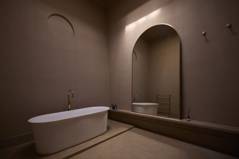A minimalist bathroom with a freestanding white tub, gold fixtures, a large arched mirror, and beige walls under soft lighting.