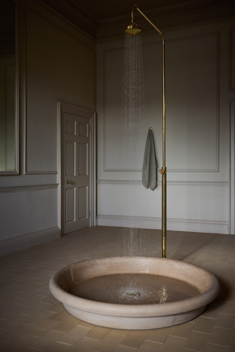 A minimalist shower with a brass fixture and beige round basin stands in an empty room with neutral walls, herringbone tile floor, and a towel draped over the shower pipe.