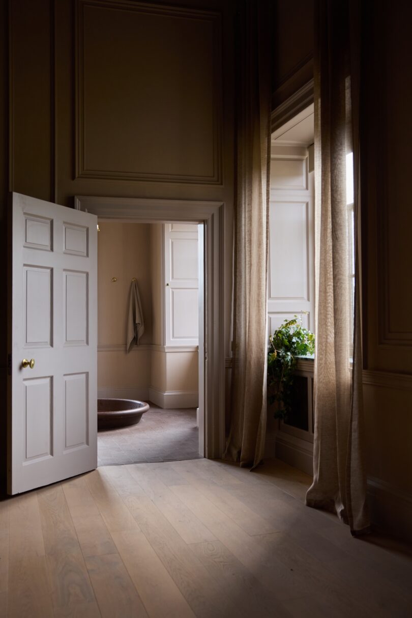 A softly lit room with wooden floors, tall windows with beige curtains, an open door, and a glimpse of a towel and basin in the adjoining space.
