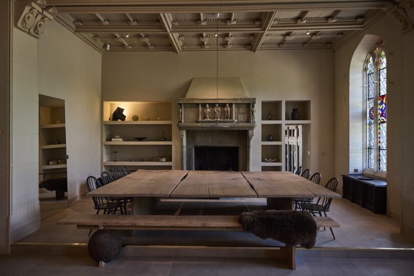 A large wooden dining table with benches is centered in a room with a stone fireplace, high ceiling, shelving, and a tall stained glass window.