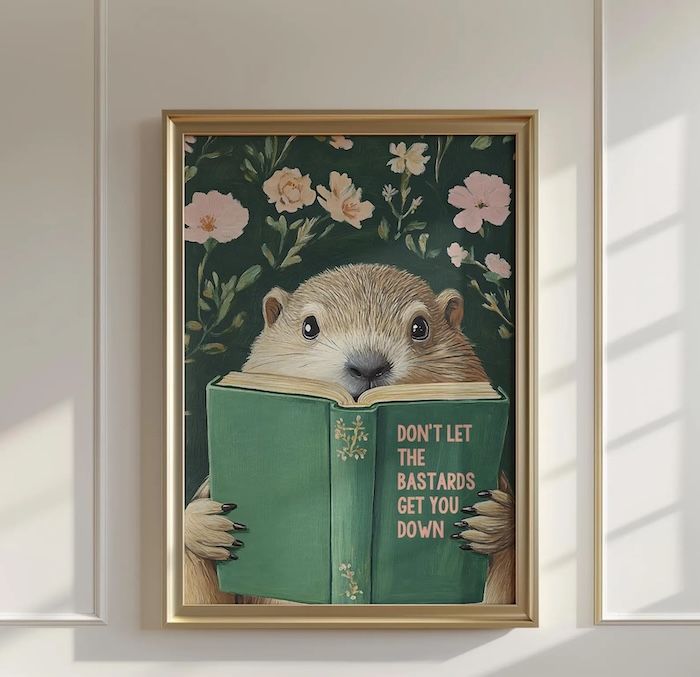 art print in a gold frame of a groundhog holding a green hardcover book with the title Don't Let the Bastards Get You Down