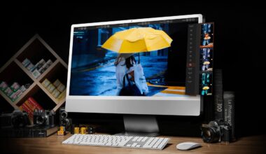 A desktop computer displays a photo of a person holding a yellow umbrella in the rain. Surrounding the monitor are film cameras, photo film rolls, photography books, and a shelf with film boxes, all set on a wooden desk.