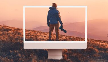 A person holding a camera stands on a grassy hill at sunset, with an oversized computer monitor frame highlighting them and the mountain landscape in the background.