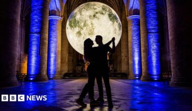 Hugh Bonneville smiles and talks as he hands a red bound hardback book back to a young girl with blonde hair, who has her back to the camera and is wearing a navy polo shirt. Bonneville is holding a black permanent marker in his other hand, and a small stack of his books lies on the wooden table in front of him.