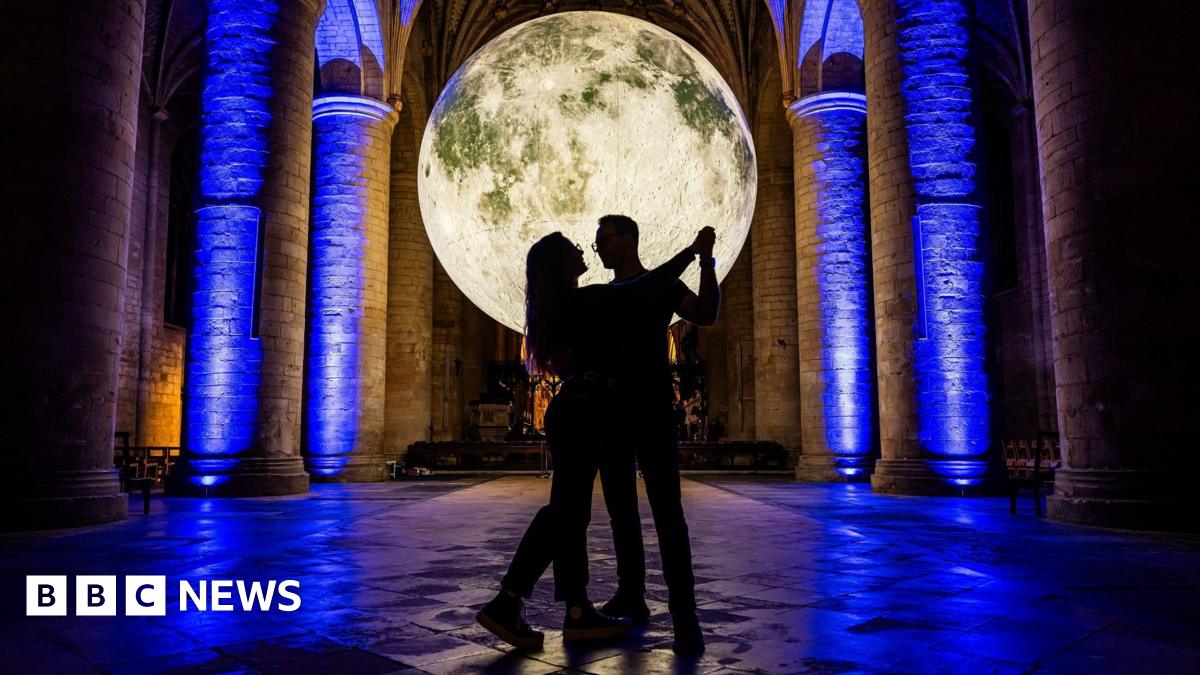 Hugh Bonneville smiles and talks as he hands a red bound hardback book back to a young girl with blonde hair, who has her back to the camera and is wearing a navy polo shirt. Bonneville is holding a black permanent marker in his other hand, and a small stack of his books lies on the wooden table in front of him.