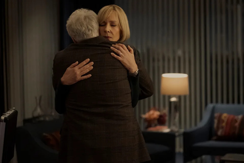 Two people embrace warmly in a softly lit room with a lamp and chairs in the background