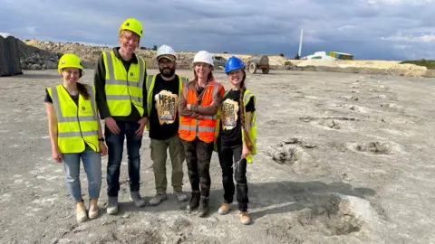 University of Birmingham Five people in yellow and orange hi-vis jackets standing in a quarry. There are sunken, fossilised dinosaur tracks in the ground that stretch into the distance