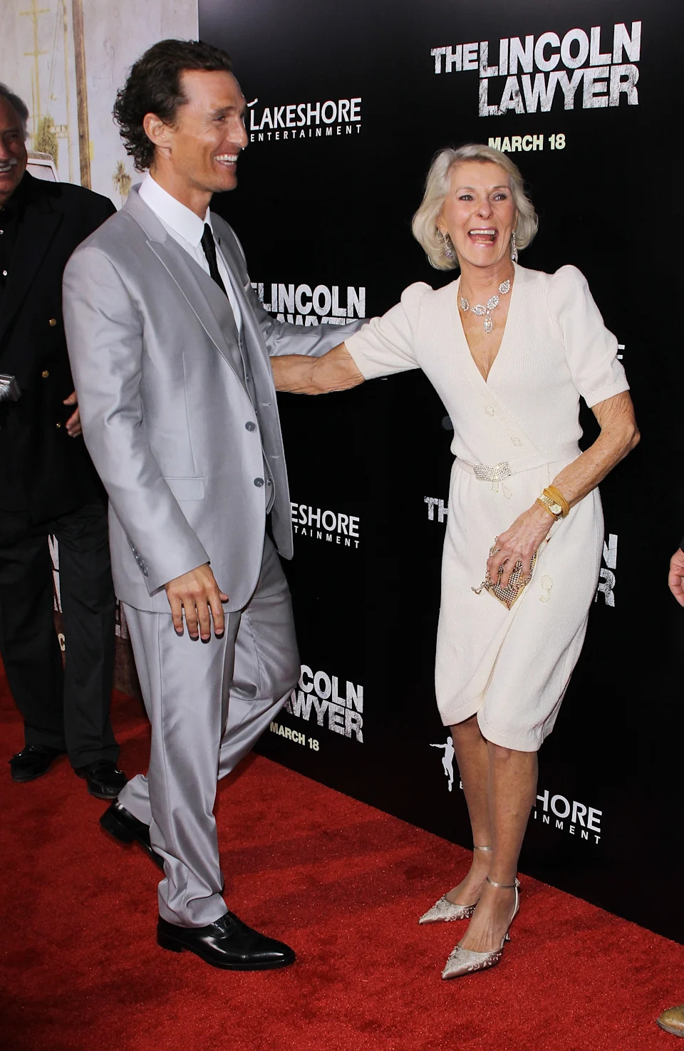 Two people smiling on a red carpet. One wears a tailored suit, the other a knee-length dress with an elegant belt. Background shows "The Lincoln Lawyer."