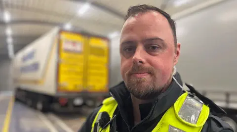 BBC Chief immigration officer Paul Harvey surveys a lorry at a Holyhead Port checkpoint. He looks at the camera, it is a head and shoulders shot. He has short, light-brown hair and a goatee. The top of his black jacket and hi-vis can be seen Behind there is a lorry that is blurred but the man is in focus. 