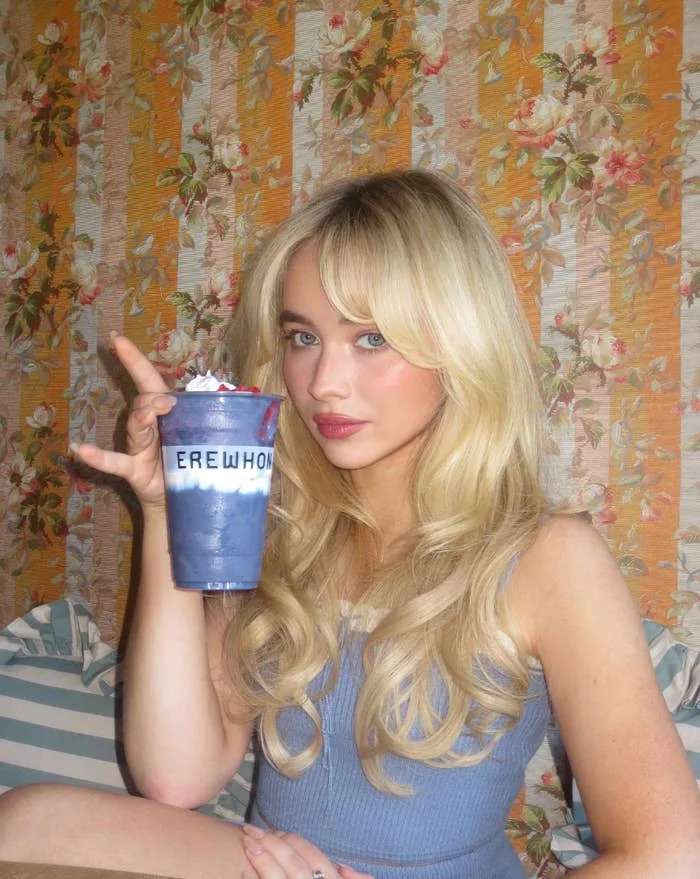 Person holding a branded cup with whipped cream, sitting indoors against a floral patterned wall, wearing a sleeveless top with wavy, long hair