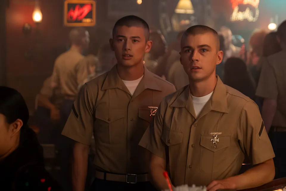 Two people in military uniforms with short haircuts stand in a crowded room, looking ahead attentively