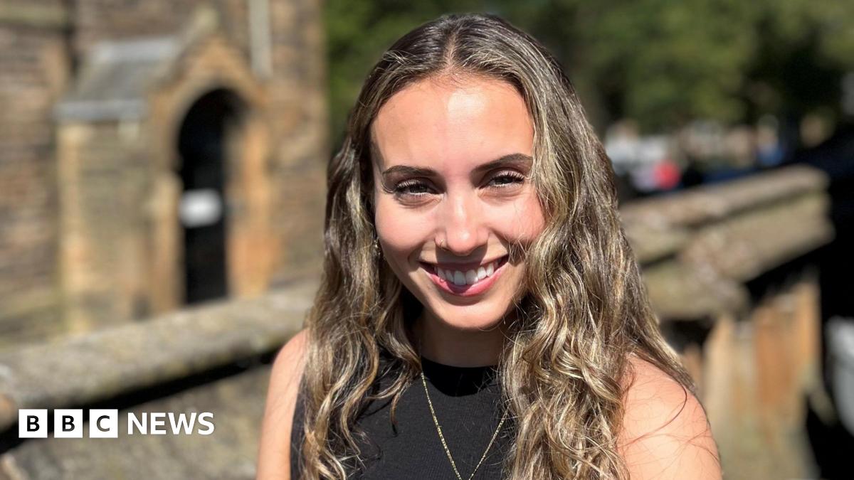 Orla Sonvico smiling at the camera. She has long brown hair and is wearing a black top. There is a wall and an old sandstone building in the distance.