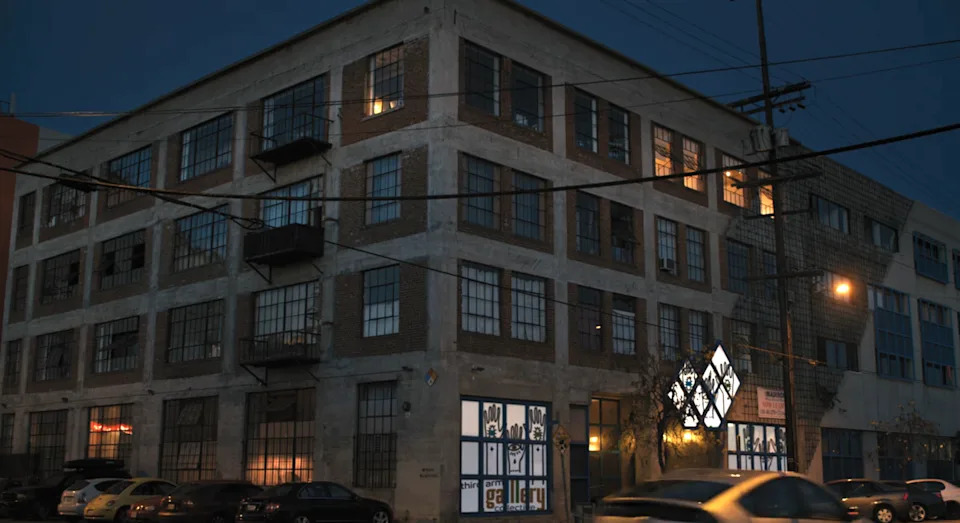 Exterior of a multi-story industrial building at dusk with illuminated signs displaying photos of individuals on the ground floor