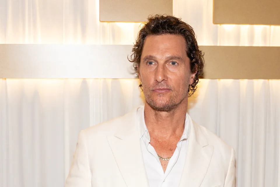 A person in a white suit and open-collar shirt is standing against a light backdrop