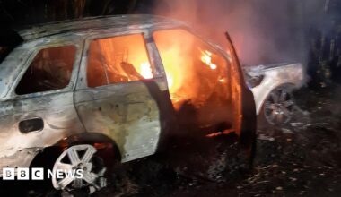 A white car with white wheel trims is engulfed in flames.