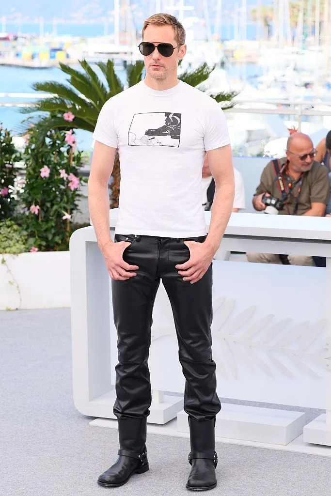 Celebrity wearing a graphic t-shirt, leather pants, and boots at an outdoor event with people and boats in the background