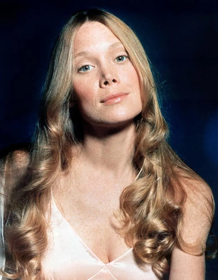 Person with long wavy hair poses against a dark background, wearing a satin top with a V-neckline