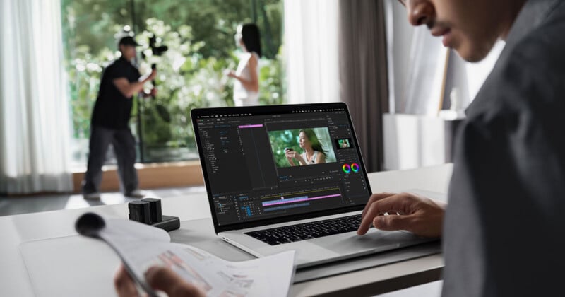 A person edits a video on a laptop at a desk, while in the background a videographer films a woman standing near large windows in a bright room.