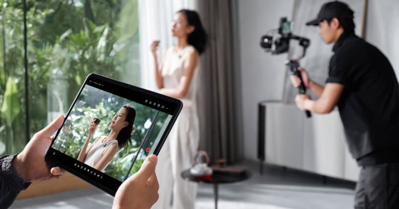 A person films a woman standing indoors while another person monitors the footage on a tablet. The room has large windows and greenery is visible outside.