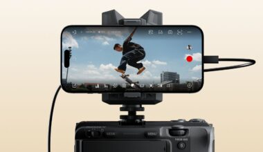 A smartphone mounted on a camera records a skateboarder mid-jump against a blue sky with clouds and distant buildings, with camera interface icons visible on the phone's screen.