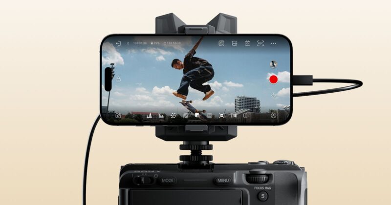 A smartphone mounted on a camera records a skateboarder mid-jump against a blue sky with clouds and distant buildings, with camera interface icons visible on the phone's screen.