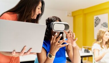 A teacher holding a laptop stands next to a student wearing virtual reality goggles, guiding her as she interacts with the device.