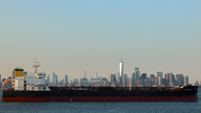 A large cargo ship leaves Port Jersey Container Terminal with the Manhattan skyline visible in the background.