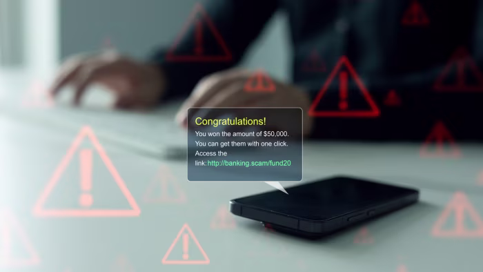 A phone displays a scam message claiming the recipient won $50,000, with warning icons superimposed and blurred hands typing in the background.