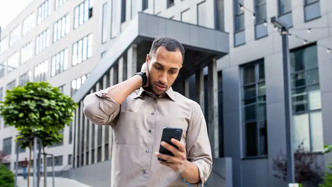 Disturbed arab man rubbing head with hand while looking at mobile phone screen on background of office building, investing myths, losing money