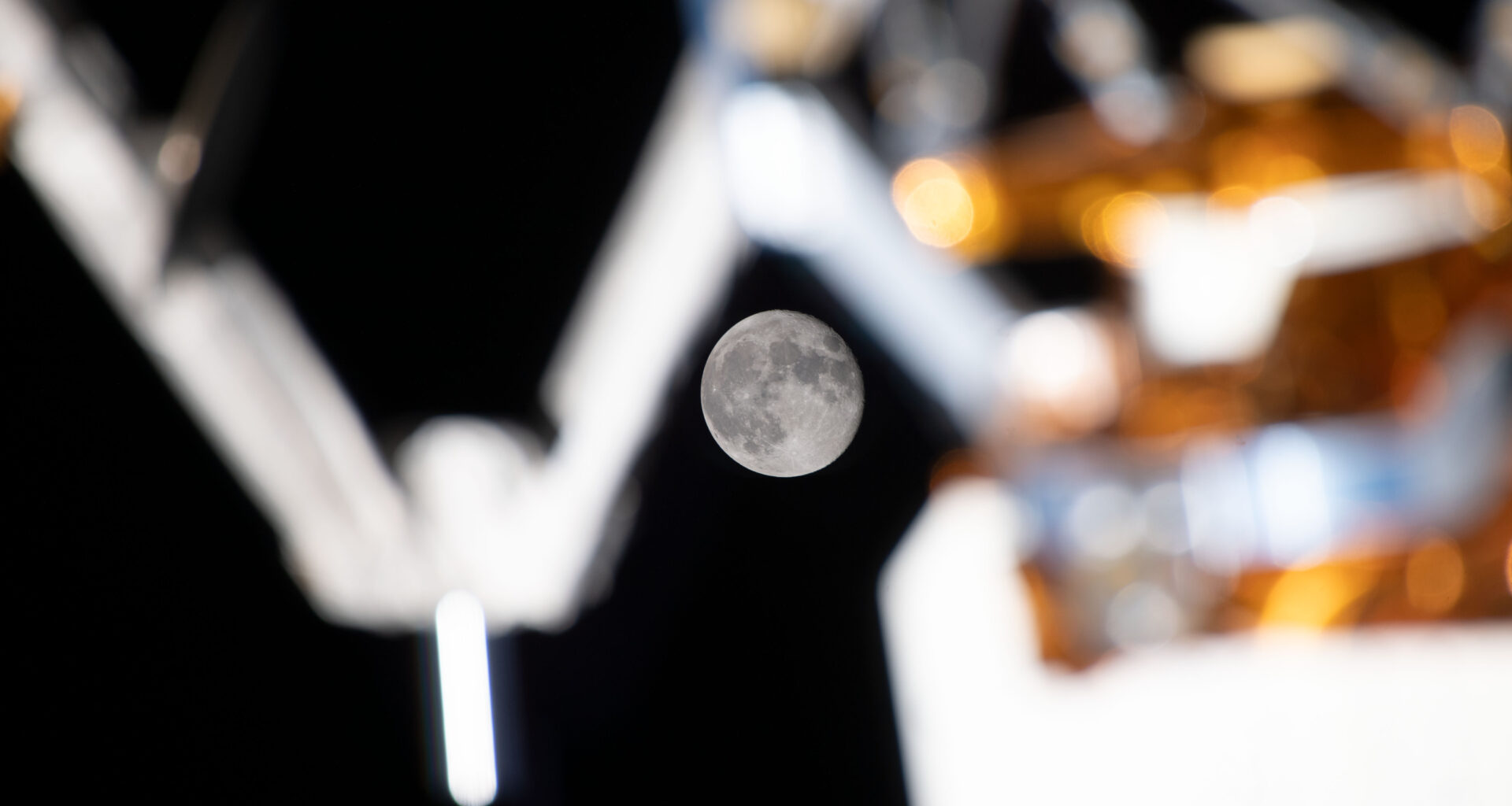 A full Moon is clearly visible in the dark background of space, framed by the blurred mechanical arms and equipment on the International Space Station’s exterior. Caption: The Moon shines between the space station’s external equipment. (Credit: NASA)