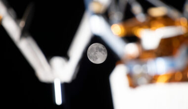 A full Moon is clearly visible in the dark background of space, framed by the blurred mechanical arms and equipment on the International Space Station’s exterior. Caption: The Moon shines between the space station’s external equipment. (Credit: NASA)