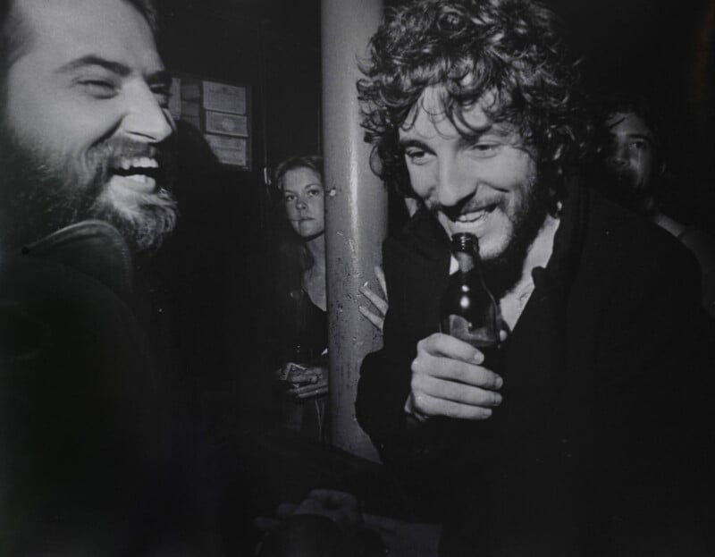 Two men laugh together at a dimly lit bar; one holds a beer bottle near his mouth. A woman and another man stand in the background, all appearing relaxed and engaged in the lively atmosphere.