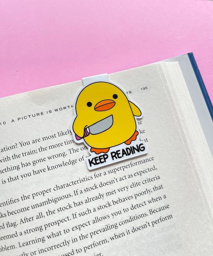 magnetic clip-on bookmark of a rond yellow duck holding a knife with text that says keep reading. the bookmark is affixed to a page in a book