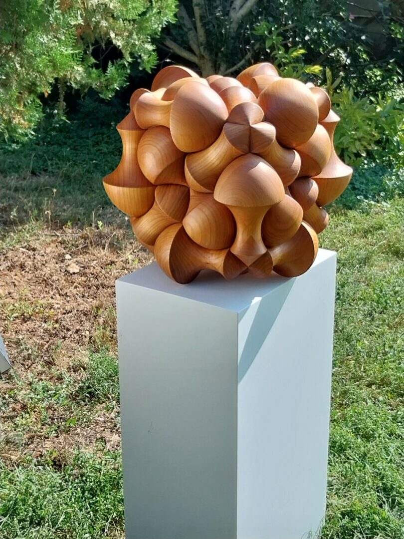 A geometric wooden sculpture composed of interlocking cone shapes sits on a white pedestal outdoors on grass, with trees in the background