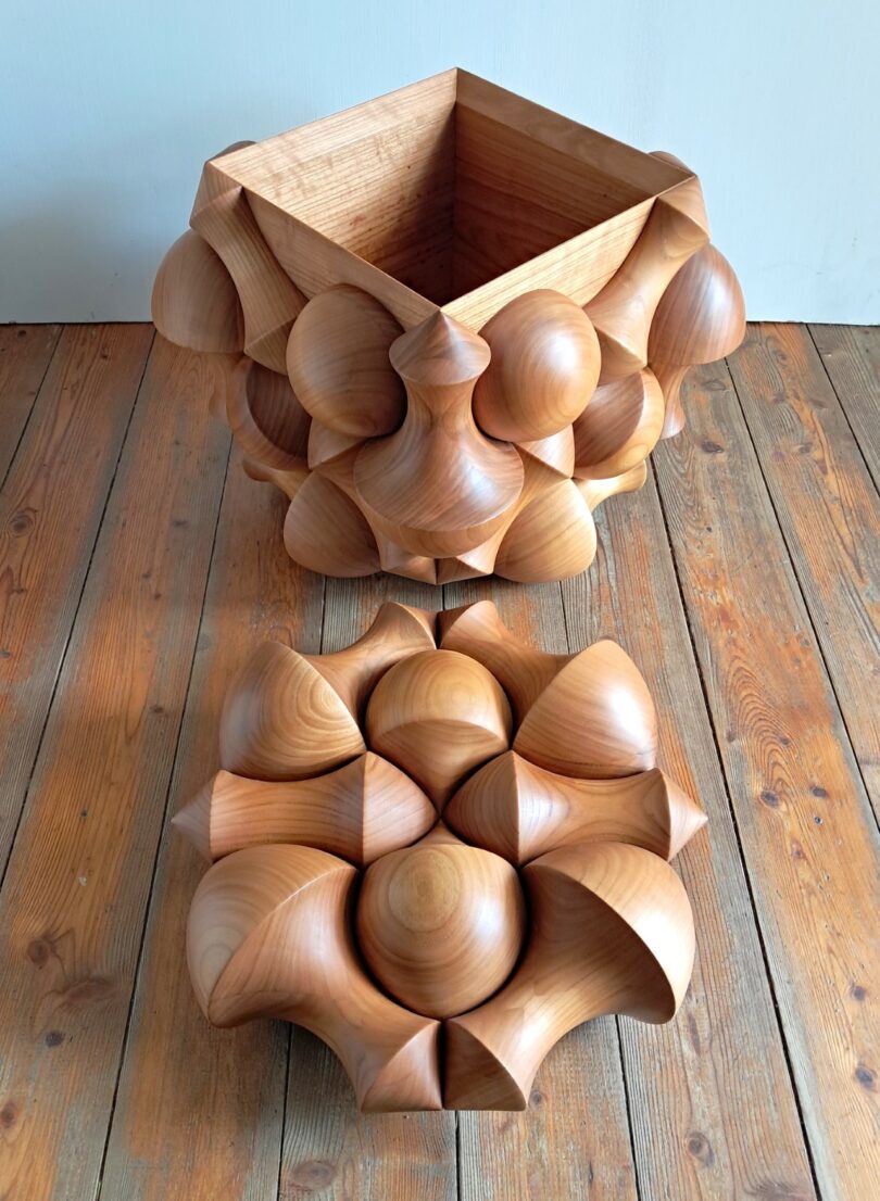 A wooden box with a geometric, sculpted exterior sits open on a wooden floor; the matching carved lid is placed nearby