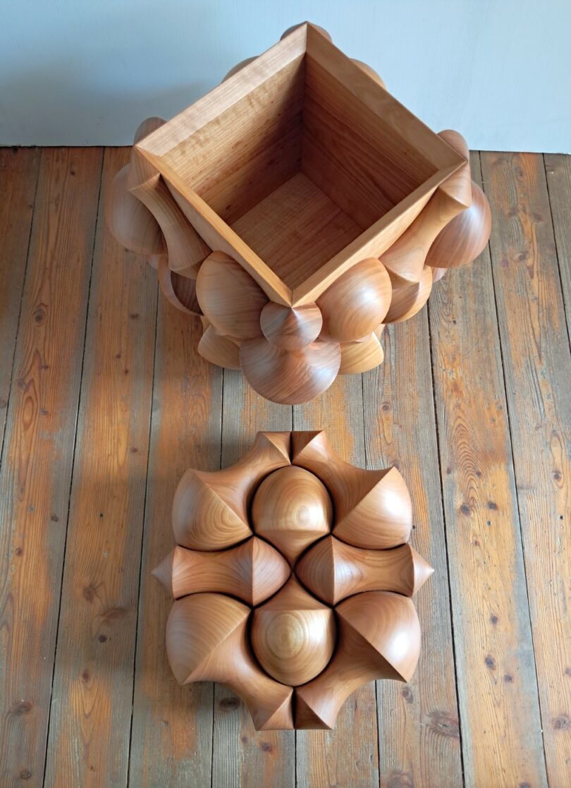 A wooden box with a geometric, sculpted exterior sits open on a wooden floor; the matching carved lid is placed nearby