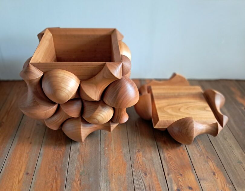 A wooden box with a geometric, rounded exterior design sits on a wooden floor; its lid is removed and placed beside it