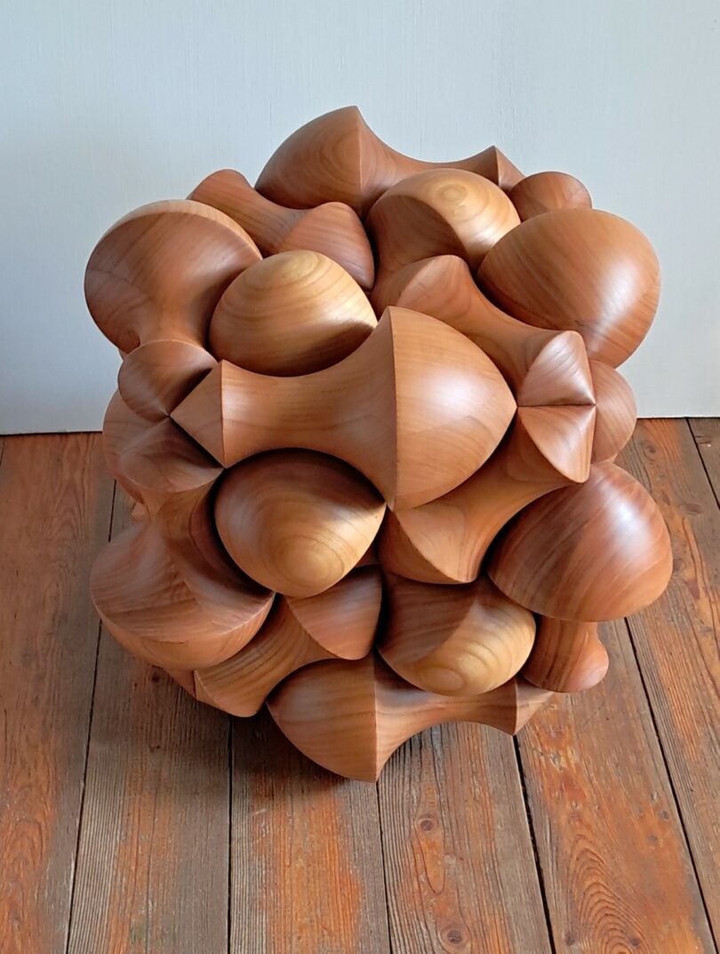 A wooden sculpture with an intricate geometric design featuring rounded, bulbous shapes, placed on a wooden floor against a plain white wall