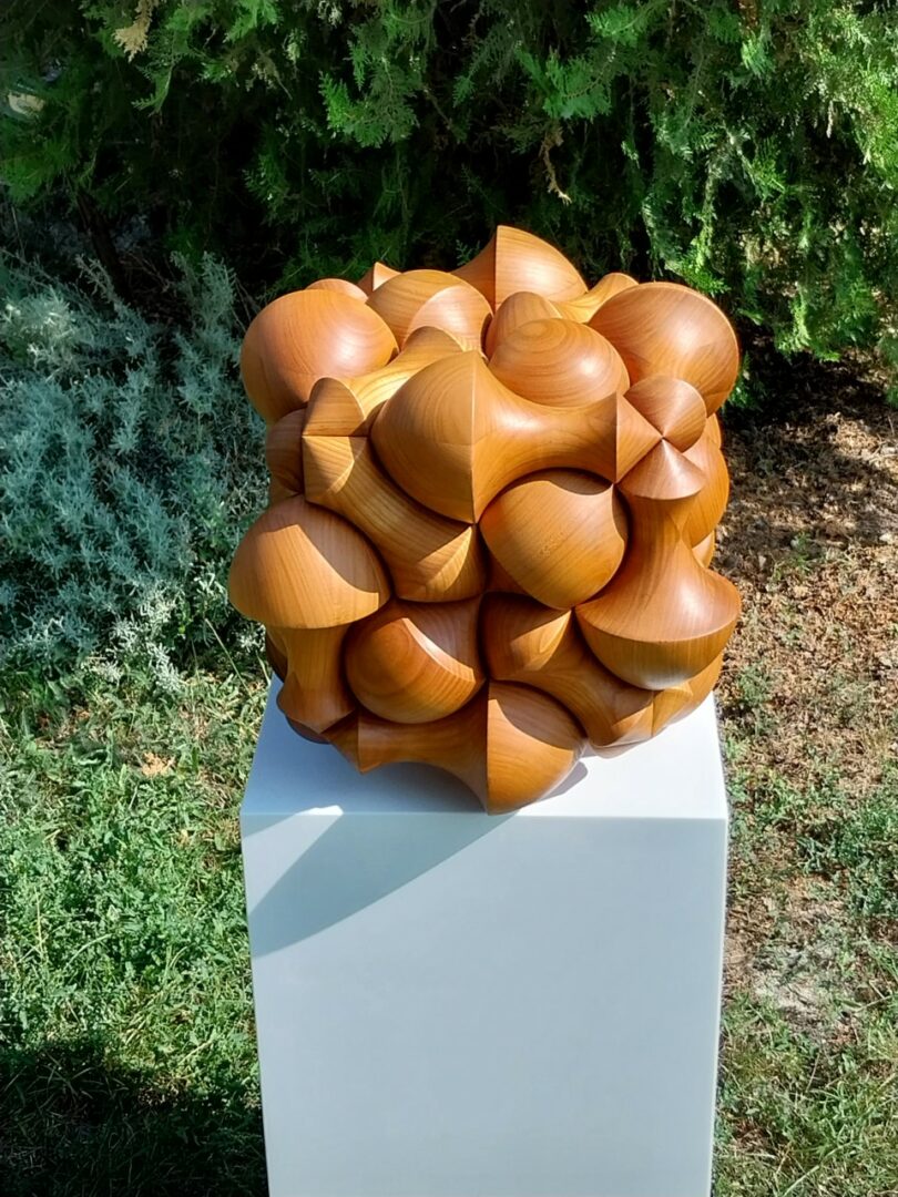 A geometric wooden sculpture composed of interlocking cone shapes sits on a white pedestal outdoors on grass, with trees in the background