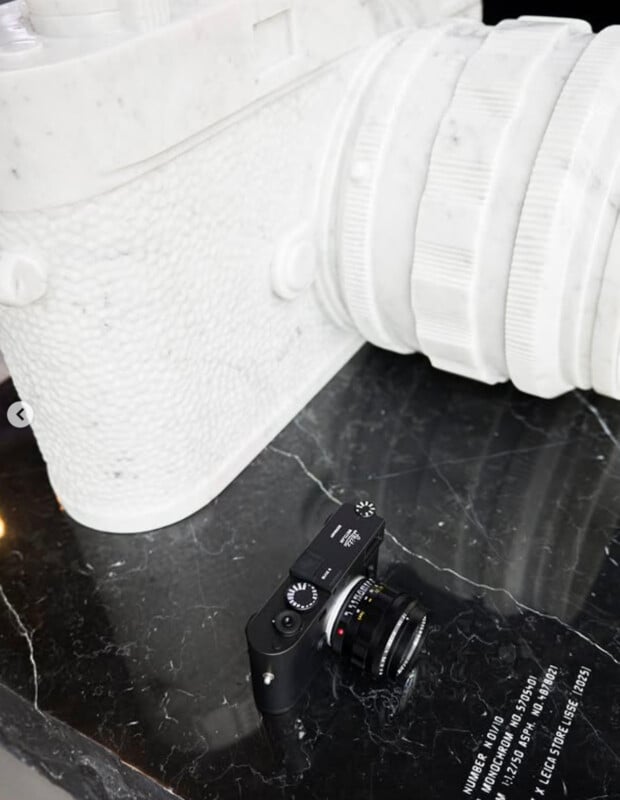 A small black Leica camera sits on a black marble surface next to a large white sculpture of a camera and lens.