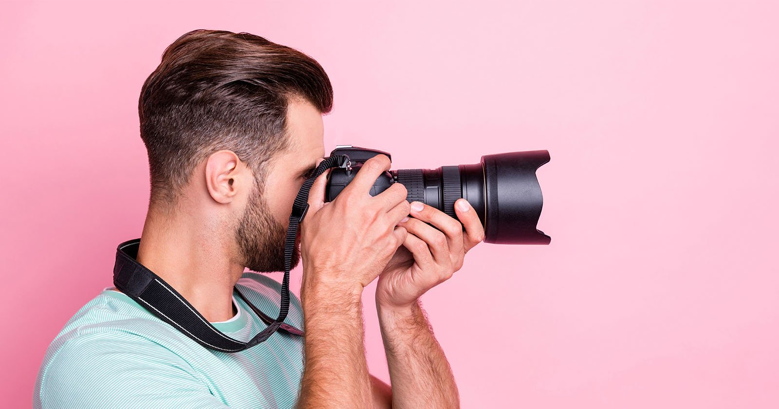 A man with short brown hair and a beard is holding a professional camera up to his face, taking a photo against a pink background. He is wearing a mint green t-shirt and has a camera strap around his neck.