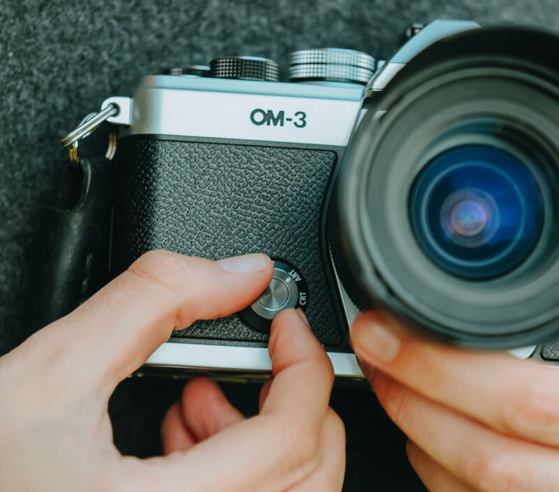 Close-up of hands adjusting a dial on an OM-3 camera, with the lens prominently in the foreground and a textured background visible.
