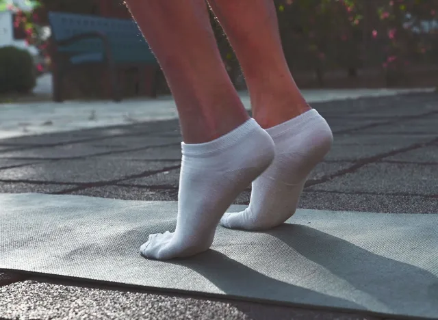 Close up of a woman's feet in white socks, warming up for a fitness workout in the park. She is standing on a yoga mat and doing calf raises.