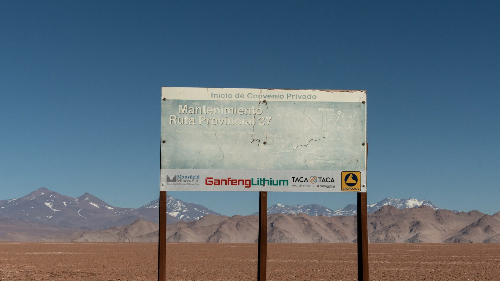 A weathered sign indicating the maintenance of Provincial Route 27, with accompanying logos of Mansfield Minera, Ganfeng Lithium, and TACA, set against a barren, mountainous landscape under a clear blue sky.